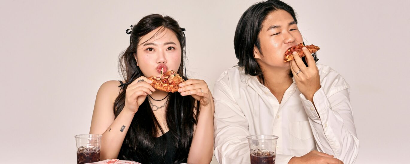 A couple savoring pizza and cola, sitting indoors with takeaway boxes.